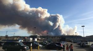 People in a parking lot with large plume of wildfire smoke in background